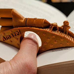 May include: A gold-colored 3D printed bookmark shaped like a rocket ship with a small astronaut figure standing on a bridge. The bookmark has the word "Amaze!" printed on it.