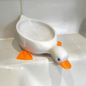 May include: A white, duck-shaped soap dish with orange feet and beak. The dish has a rounded basin for holding soap and is designed to sit on a countertop. The duck's eyes are black.
