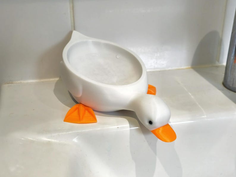 May include: A white, duck-shaped soap dish with orange feet and beak. The dish has a rounded basin for holding soap and is designed to sit on a countertop. The duck's eyes are black.