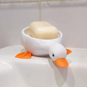 May include: A white ceramic soap dish shaped like a duck, with orange feet and beak, and black eyes. The dish holds a bar of light-colored soap. The duck-shaped dish is a decorative bathroom accessory.