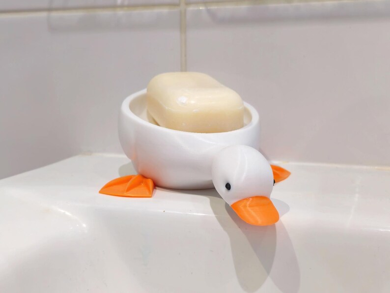 May include: A white ceramic soap dish shaped like a duck, with orange feet and beak, and black eyes. The dish holds a bar of light-colored soap. The duck-shaped dish is a decorative bathroom accessory.
