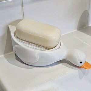 May include: A white duck-shaped soap dish with an orange beak and black eyes, holding a bar of soap. The soap dish has a honeycomb-patterned surface to allow for drainage. The soap is a light beige color.