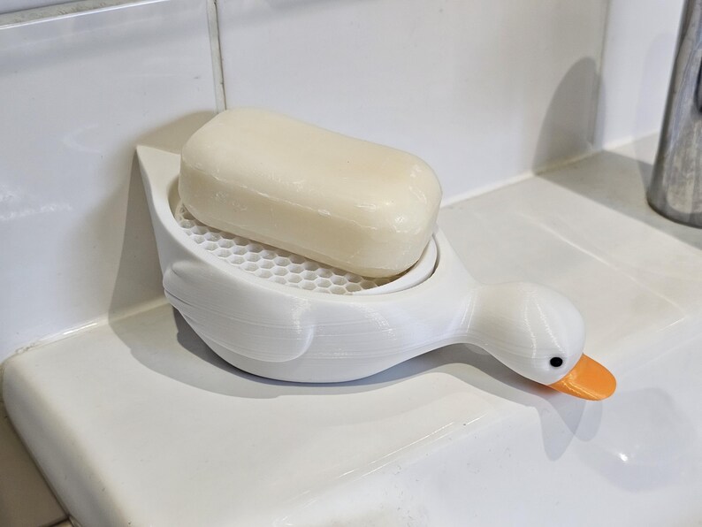 May include: A white duck-shaped soap dish with an orange beak and black eyes, holding a bar of soap. The soap dish has a honeycomb-patterned surface to allow for drainage. The soap is a light beige color.