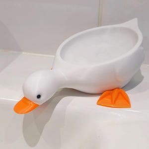 May include: A white, duck-shaped soap dish with an orange beak and feet. The dish is designed to hold soap and has a simple, modern aesthetic. The duck's head is lowered, and its feet are positioned on the surface.