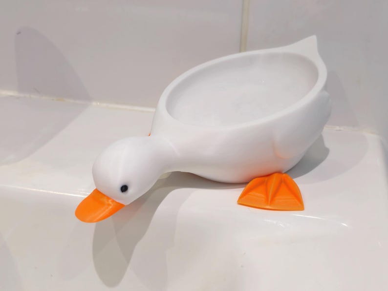 May include: A white, duck-shaped soap dish with an orange beak and feet. The dish is designed to hold soap and has a simple, modern aesthetic. The duck's head is lowered, and its feet are positioned on the surface.