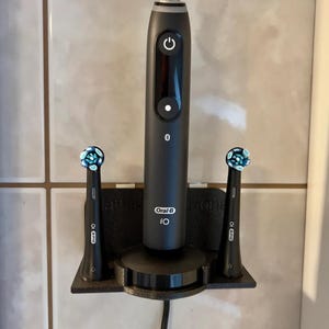May include: A black wall-mounted toothbrush holder with two Oral-B iO electric toothbrushes and a charging station. The holder has a black base and two slots for the toothbrushes. The toothbrushes are black with blue and white accents.