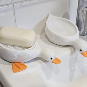 May include: Two white duck-shaped soap dishes with orange beaks and feet. One holds a bar of soap. The soap dishes are on a white surface, next to a chrome faucet. The soap dish has a honeycomb-patterned base.