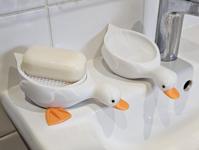 May include: Two white duck-shaped soap dishes with orange beaks and feet. One holds a bar of soap. The soap dishes are on a white surface, next to a chrome faucet. The soap dish has a honeycomb-patterned base.