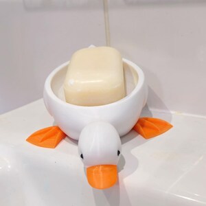 May include: A white ceramic duck-shaped soap dish with orange feet and beak. A bar of light-colored soap rests inside the dish. The soap dish is on a white surface.