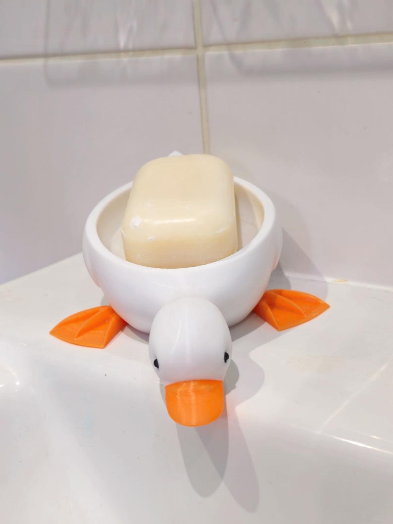 May include: A white ceramic duck-shaped soap dish with orange feet and beak. A bar of light-colored soap rests inside the dish. The soap dish is on a white surface.