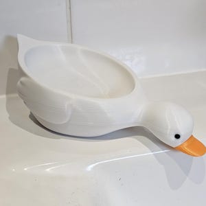 May include: A white, duck-shaped soap dish with an orange beak and black eyes. The dish is made of a smooth material and has a curved design. The duck is positioned on a white surface, suggesting a bathroom or kitchen setting.