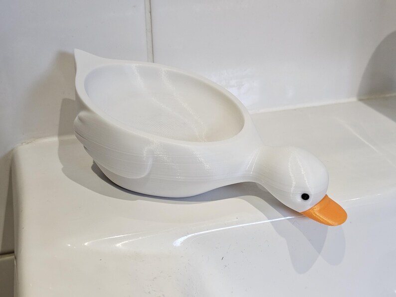 May include: A white, duck-shaped soap dish with an orange beak and black eyes. The dish is made of a smooth material and has a curved design. The duck is positioned on a white surface, suggesting a bathroom or kitchen setting.