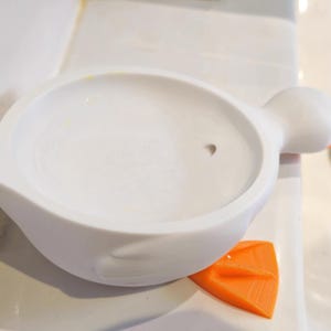 May include: A white, duck-shaped soap dish with an orange foot and an orange eye. The dish has a round, shallow basin. The duck's head is on the right side of the dish.