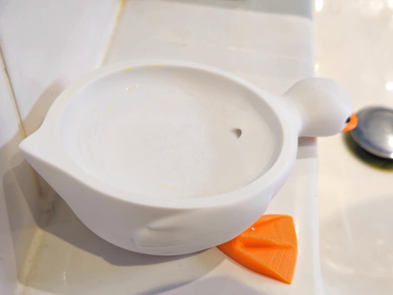 May include: A white, duck-shaped soap dish with an orange foot and an orange eye. The dish has a round, shallow basin. The duck's head is on the right side of the dish.