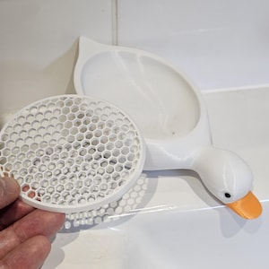 May include: A white, 3D-printed goose-shaped soap dish with an orange beak and black eyes. A separate, round, white soap dish with a honeycomb pattern is next to the goose. The soap dish is designed to hold soap.