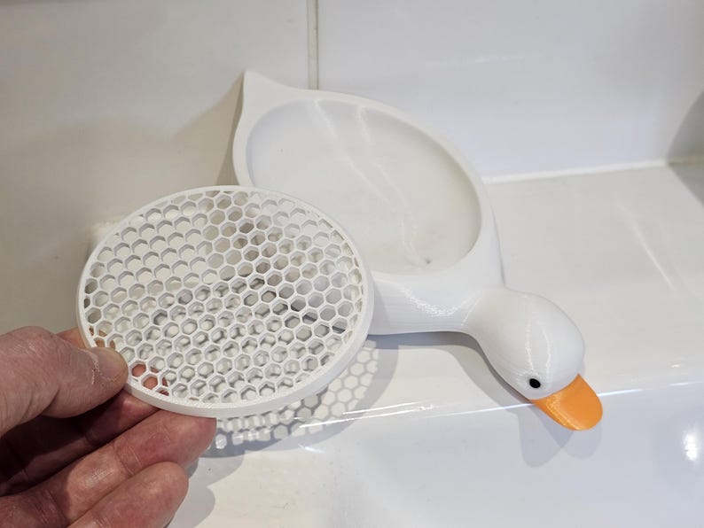 May include: A white, 3D-printed goose-shaped soap dish with an orange beak and black eyes. A separate, round, white soap dish with a honeycomb pattern is next to the goose. The soap dish is designed to hold soap.