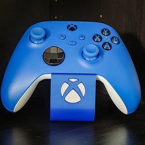 May include: A blue Xbox controller on a blue stand. The controller has black buttons and white detailing. The stand displays the Xbox logo in silver and white.