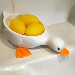 May include: A white duck-shaped soap dish holding three yellow oval soaps. The duck has an orange beak and feet, with a black eye. The dish is on a white surface.