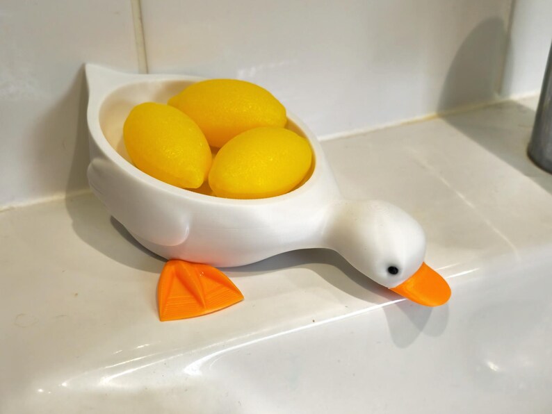 May include: A white duck-shaped soap dish holding three yellow oval soaps. The duck has an orange beak and feet, with a black eye. The dish is on a white surface.