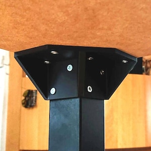 May include: A black metal table leg supporting a brown tabletop. The leg has a square base and a triangular bracket connecting to the tabletop. Screws secure the bracket to both the leg and the tabletop.