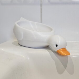 May include: A white duck-shaped container with an orange beak and black eyes. The container is designed to hold small items. The duck is resting on a white surface.