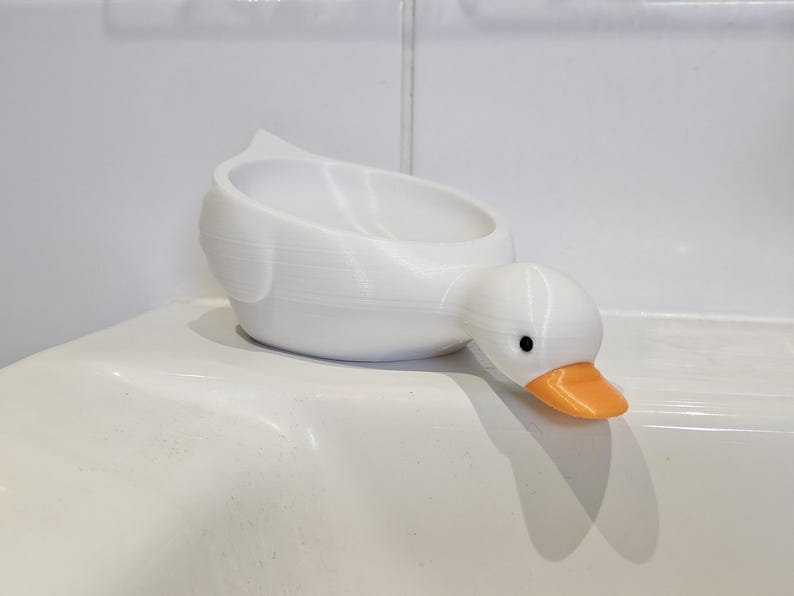 May include: A white duck-shaped container with an orange beak and black eyes. The container is designed to hold small items. The duck is resting on a white surface.