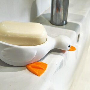May include: A white duck-shaped soap dish with an orange beak and feet holds a bar of soap. The soap dish is on a white surface, near a faucet. The soap is a light beige color.