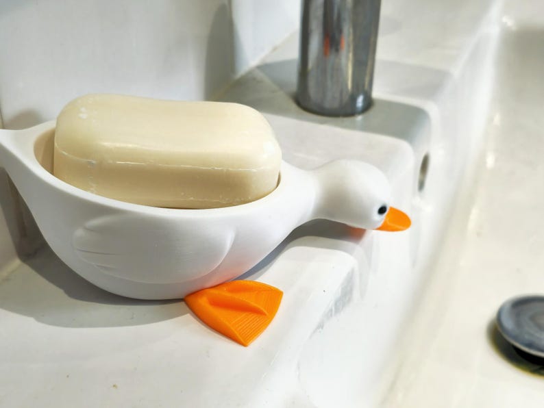 May include: A white duck-shaped soap dish with an orange beak and feet holds a bar of soap. The soap dish is on a white surface, near a faucet. The soap is a light beige color.