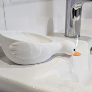 May include: A white, duck-shaped soap dish with an orange beak and black eyes. The dish is resting on a white sink, near a chrome faucet. The dish is designed to hold soap and adds a whimsical touch to a bathroom.
