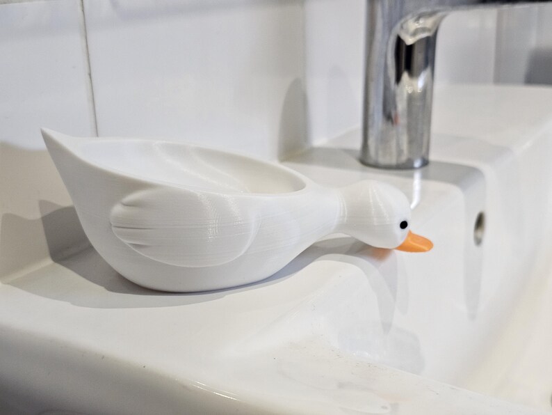 May include: A white, duck-shaped soap dish with an orange beak and black eyes. The dish is resting on a white sink, near a chrome faucet. The dish is designed to hold soap and adds a whimsical touch to a bathroom.