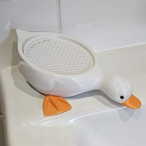 May include: A white duck-shaped soap dish with an orange beak and feet. The dish has a textured, honeycomb-patterned surface for holding soap. The duck is resting on a white surface.