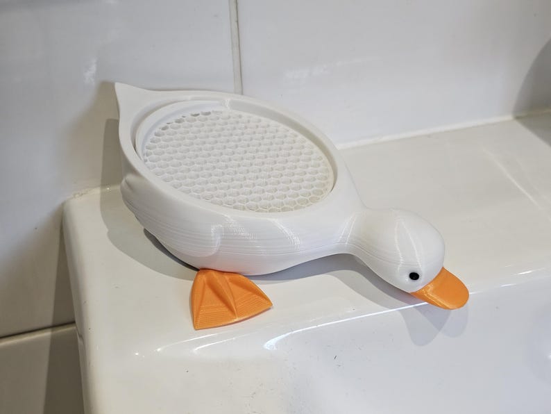 May include: A white duck-shaped soap dish with an orange beak and feet. The dish has a textured, honeycomb-patterned surface for holding soap. The duck is resting on a white surface.