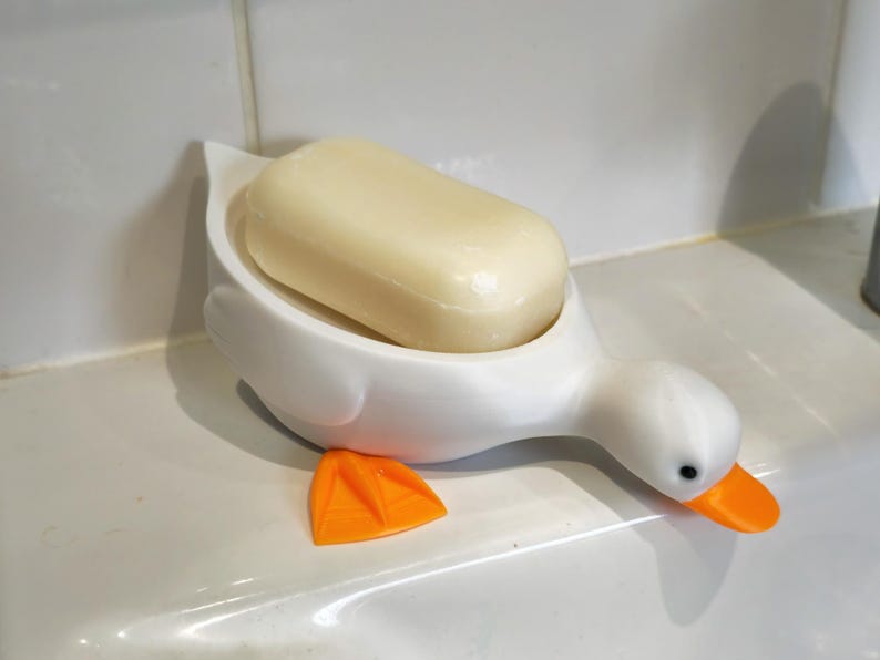May include: A white duck-shaped soap dish with an orange beak and feet, holding a bar of light yellow soap. The soap dish is on a white surface, likely in a bathroom setting.