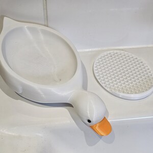 May include: A white, 3D-printed soap dish in the shape of a goose, with an orange beak and a separate, round, textured soap rest. The dish is designed to hold soap and adds a decorative touch to a bathroom.