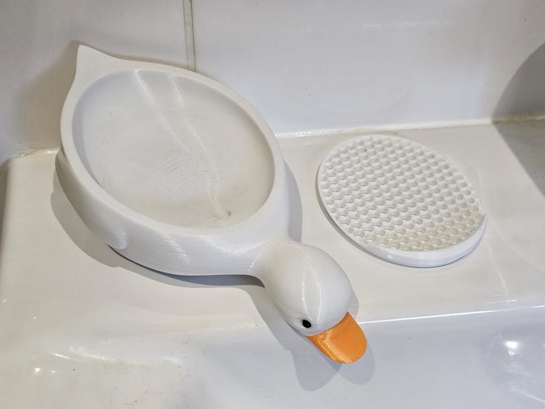 May include: A white, 3D-printed soap dish in the shape of a goose, with an orange beak and a separate, round, textured soap rest. The dish is designed to hold soap and adds a decorative touch to a bathroom.