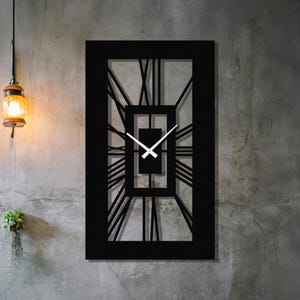 May include: A black rectangular wall clock with Roman numerals. The clock has a geometric design with a central square and radiating lines. The clock hands are white. The clock is mounted on a gray concrete wall.