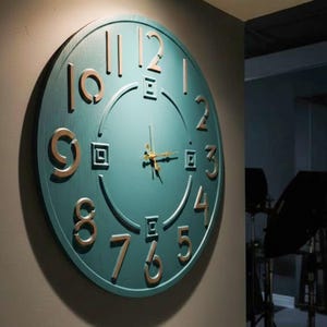 Può includere: Un grande orologio da parete rotondo con un quadrante turchese e numeri color oro. Le lancette dell'orologio sono dorate e i numeri sono in un font moderno. L'orologio è montato su una parete grigia e il design generale è contemporaneo.