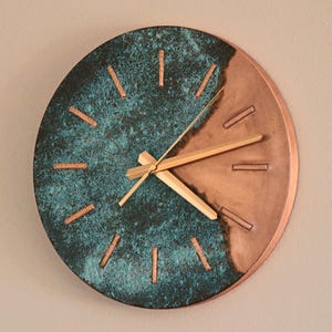 Könnte beinhalten: Runde Wanduhr mit zweifarbigem Design. Die linke Seite hat eine strukturierte türkisfarbene Oberfläche, während die rechte Seite kupferfarben ist. Die Uhr hat goldfarbene Zeiger und kupferfarbene Stundenmarkierungen. Ein dekoratives Stück.