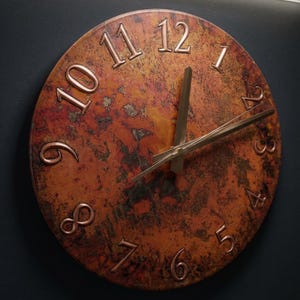 Può includere: Un orologio da parete rotondo con un quadrante color rame e numeri in bronzo in rilievo. Le lancette dell'orologio sono in bronzo. Il quadrante ha un aspetto screziato e strutturato con sfumature di arancione, marrone e nero. L'orologio è montato su uno sfondo scuro.