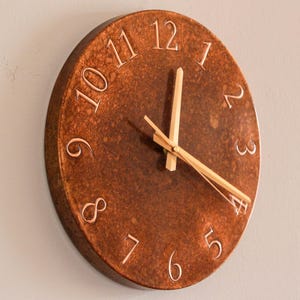 May include: A round, copper-colored wall clock with raised numerals and hands. The clock face has a textured finish, and the hands are made of light-colored wood. The numerals are in a classic font, and the clock is mounted on a neutral-colored wall.