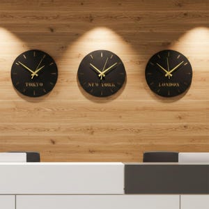 Peut inclure: Trois horloges rondes noires avec des aiguilles dorées et des noms de villes : Tokyo, New York et Londres. Elles sont fixées sur un mur en bois brun clair. Les horloges ont un design minimaliste.