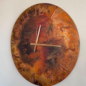 May include: A round wall clock with a textured, mottled surface in shades of copper, bronze, and dark brown. The clock face features raised, embossed numerals from 1 to 12 in a metallic finish, paired with thin, straight gold-toned clock hands.