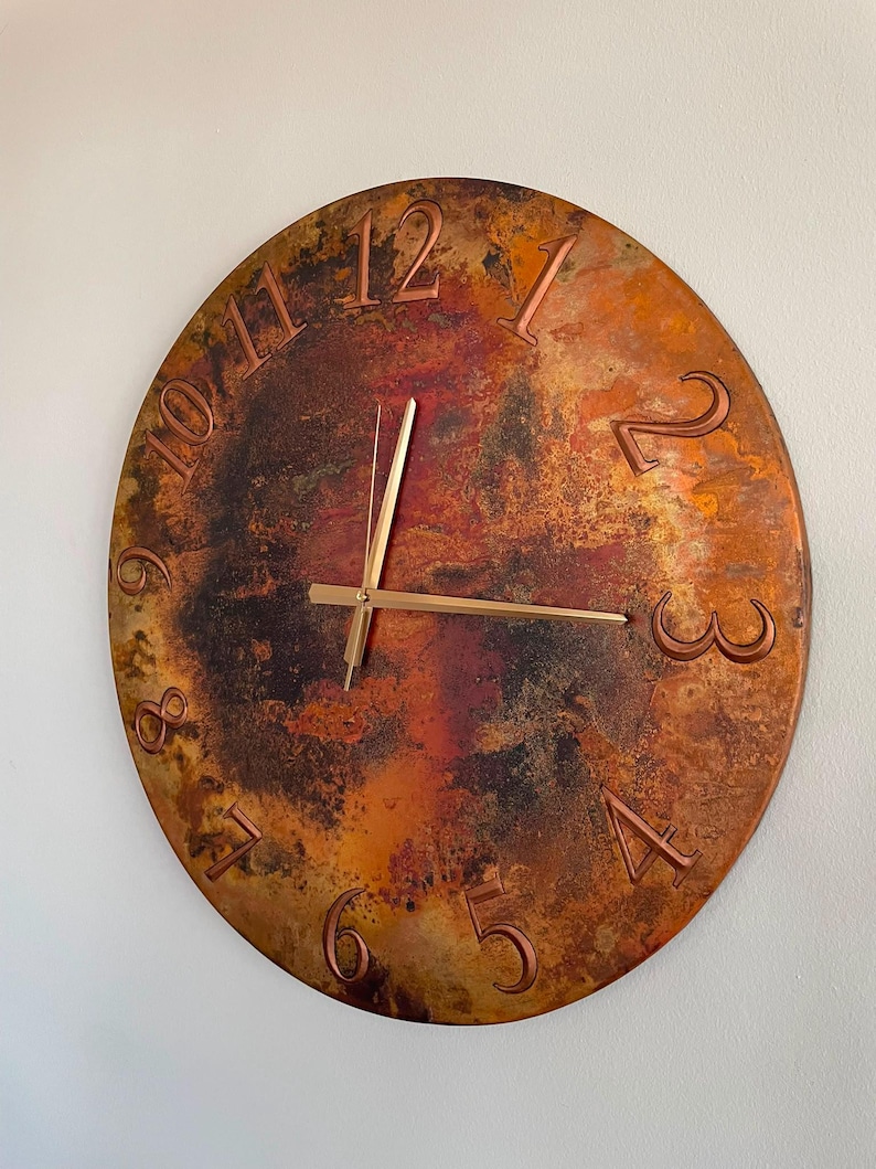 May include: A round wall clock with a textured, mottled surface in shades of copper, bronze, and dark brown. The clock face features raised, embossed numerals from 1 to 12 in a metallic finish, paired with thin, straight gold-toned clock hands.
