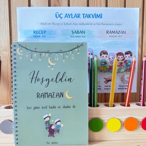 Könnte beinhalten: Ein hellgrünes Spiralnotizbuch mit den Worten "Hoşgeldin RAMAZAN" und einer Mond-Illustration. Ein Kalender mit türkischem Text befindet sich hinter dem Notizbuch, zusammen mit Buntstiften in einem Holzhalter.