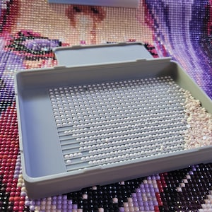 May include: A gray plastic tray with a grid pattern, partially filled with white and beige diamond painting beads. The tray sits on a partially completed diamond painting with a colorful, abstract design.
