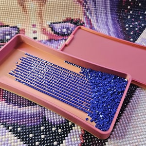 May include: A pink diamond painting tray with a lid, filled with blue diamond-shaped rhinestones. The tray has a series of grooves for organizing the gems. The background shows a partially completed diamond painting.