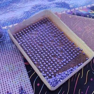 May include: A clear plastic storage jar containing small purple square diamond painting drills, next to a rectangular white plastic tray filled with rows of identical purple square drills, positioned on a printed adhesive canvas sheet.