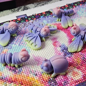 May include: Five small, articulated insect-like figures in a gradient of purple, pink, and light green. The figures include two winged insects, one caterpillar-like creature, and two rounded beetle-like forms, all featuring a matte, textured finish.