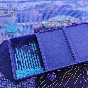 May include: A purple plastic sorting tray with three rectangular compartments containing small, square, light blue diamond painting beads. A clear plastic storage jar with a label reading 55 P 3846 sits nearby, filled with matching light blue beads.