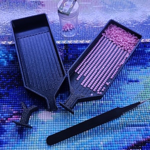 May include: Two black plastic diamond painting trays, one empty and one filled with small pink square resin drills. A clear plastic storage vial containing pink drills, a black plastic stopper, and a pair of black metal tweezers are included.